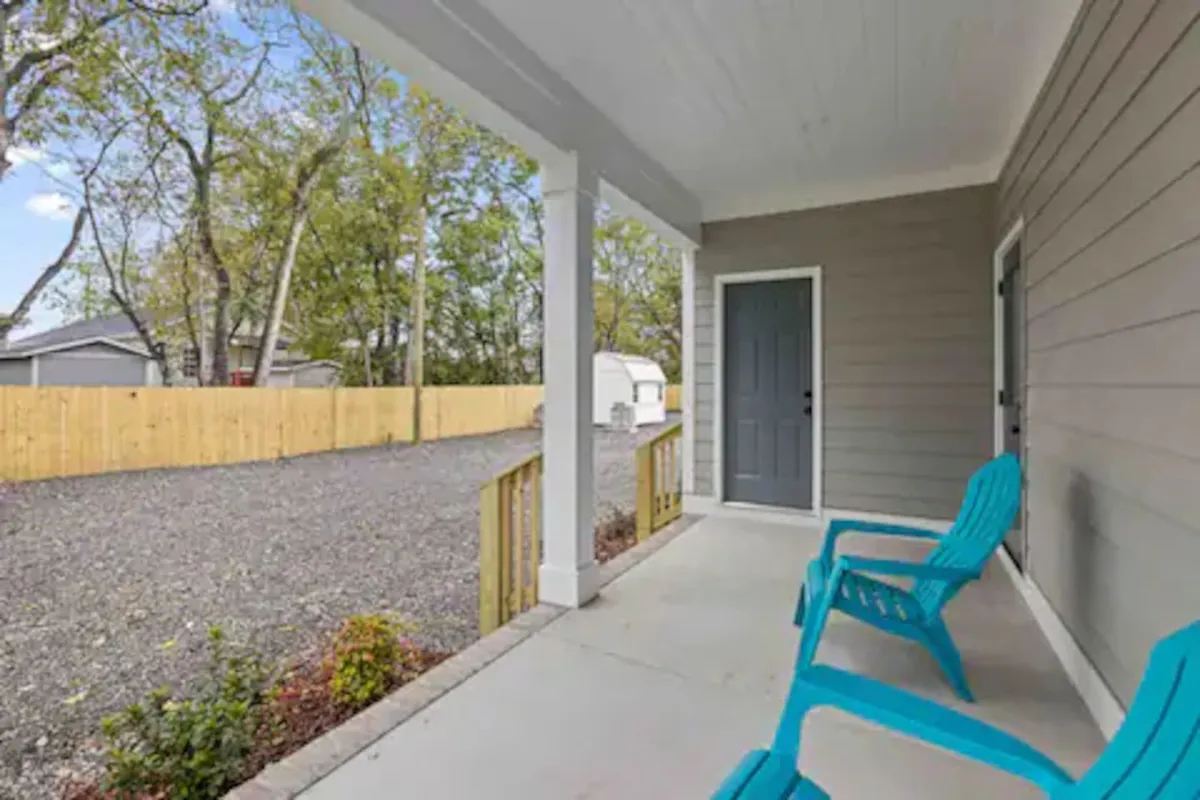 An exterior shot featuring two turquoise chairs by a grey door, a fenced yard, and a gravel driveway. The scene is tranquil yet practical, suitable for arrivals and outdoor gatherings, while highlighting available parking and ease of access.