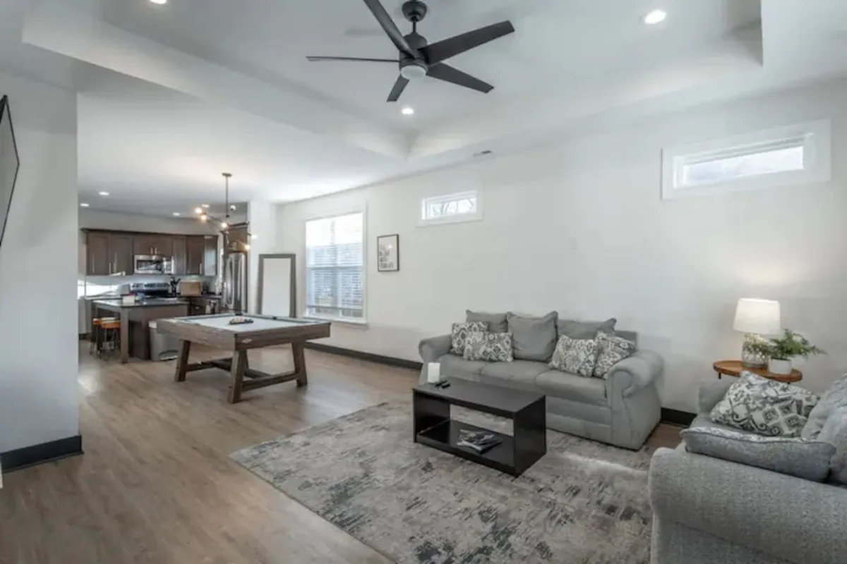 A bright, open living room featuring a plush sofa adorned with decorative pillows, a coffee table at the center, and a clear view into a spacious dining area in the background. The blend of seating and dining elements demonstrates how both living and dining functions coexist comfortably in one hub.