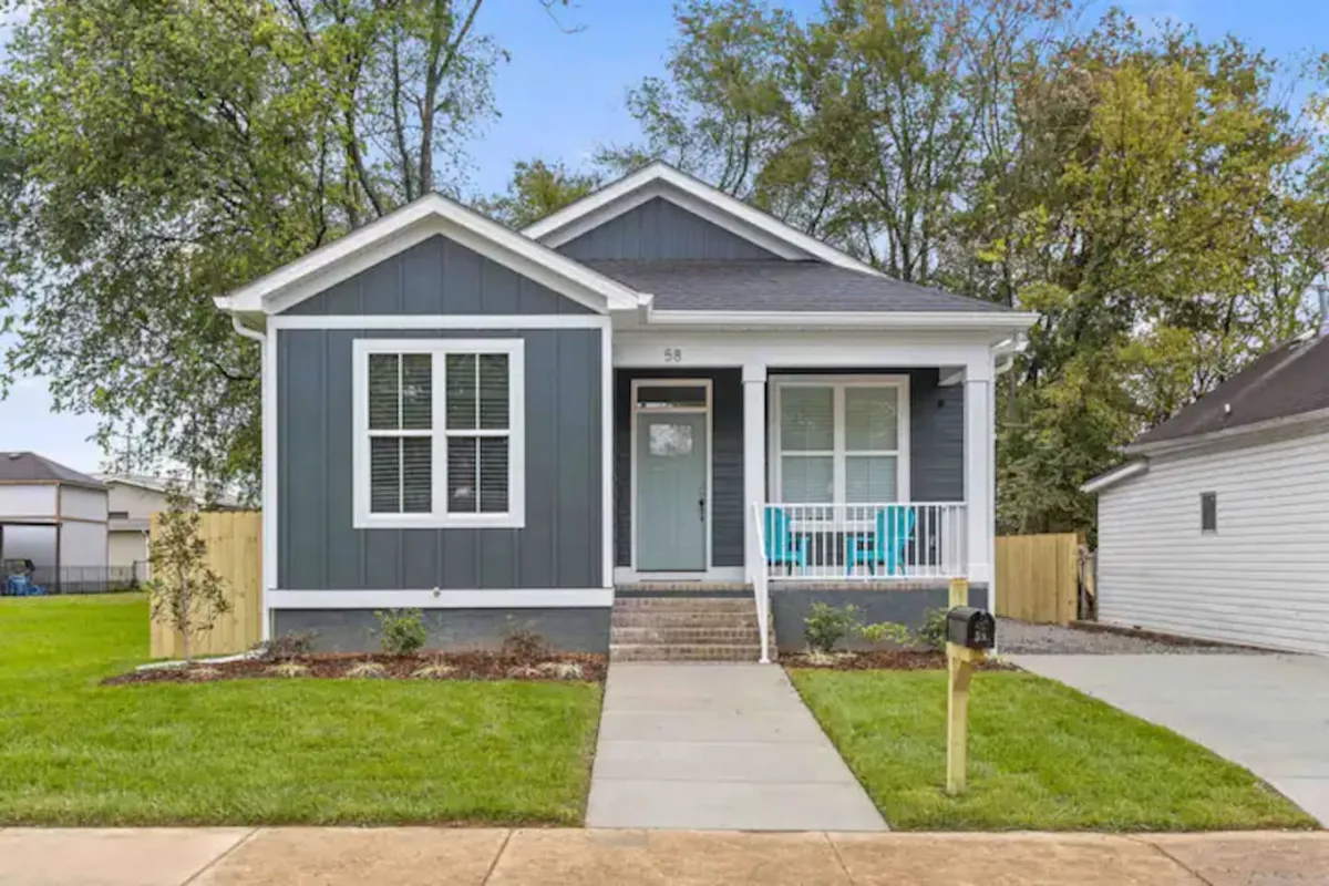 A welcoming front exterior view shows a landscaped garden, shuttered windows, a modern entrance, and a wide yard where children can play. The front porch is furnished for relaxation, and the overall mood is friendly and inviting.