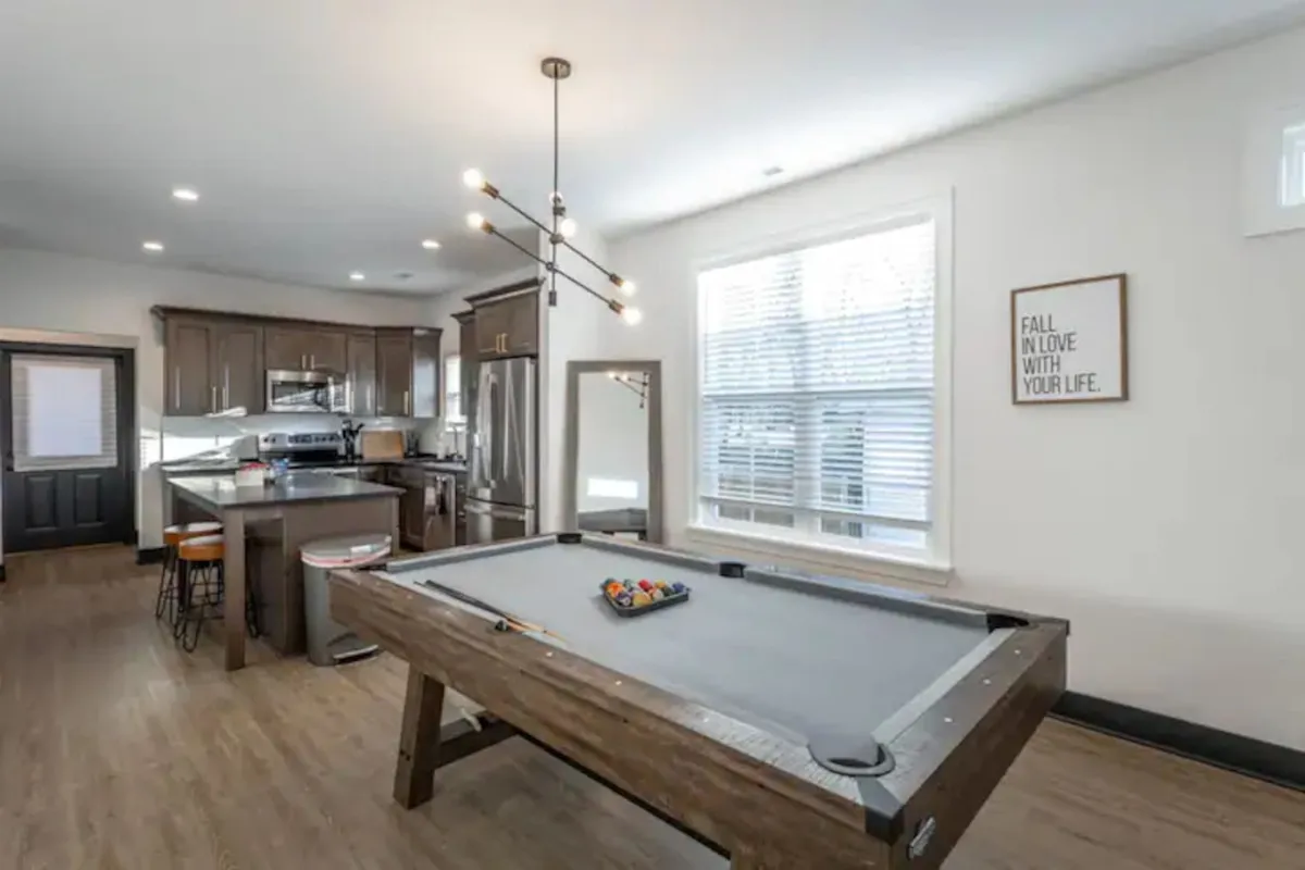 A spacious interior featuring a pool table, kitchen island, large windows, and framed wall art. The room is designed for both socializing and relaxing, showcasing multiple gathering areas ideal for large groups.
