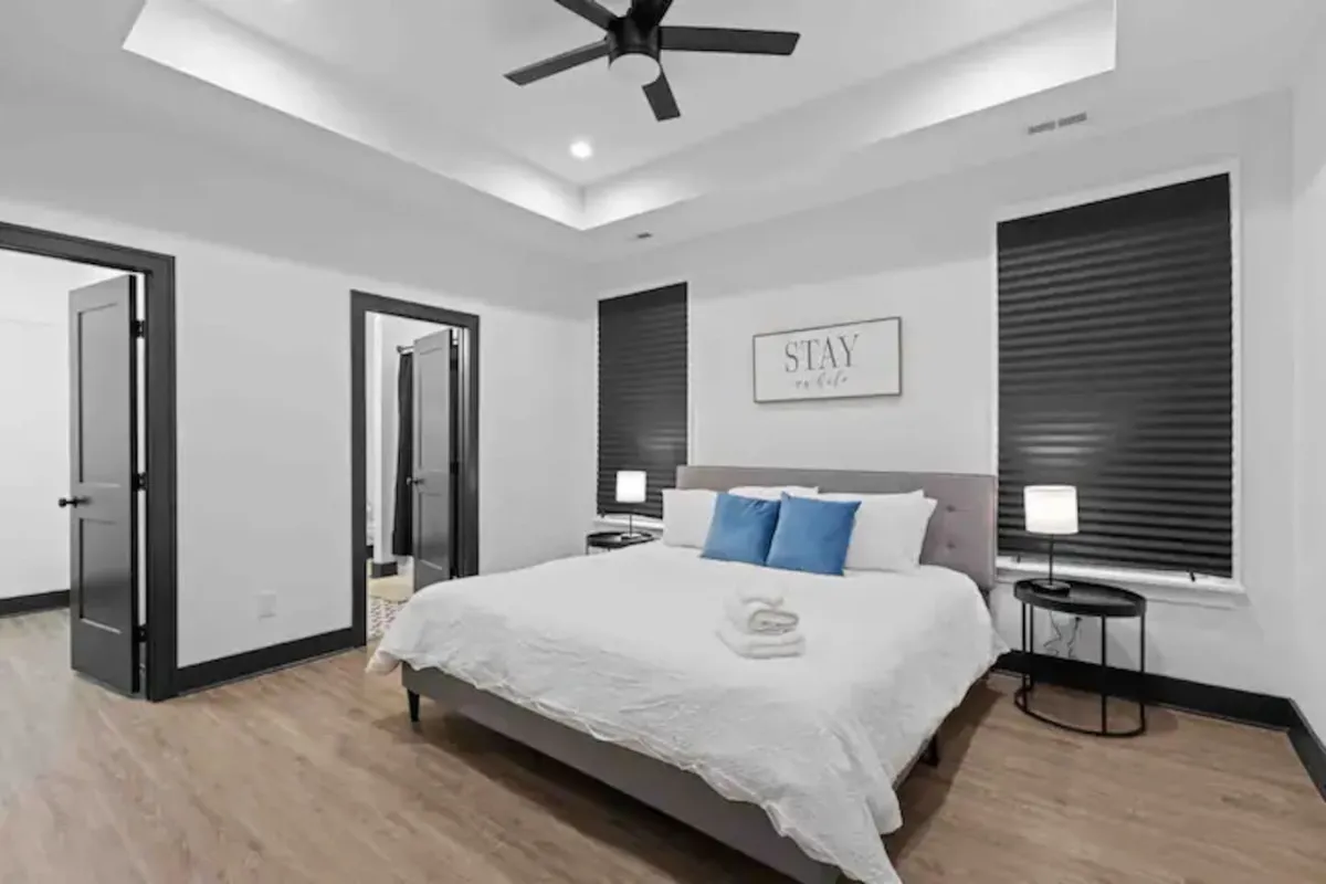 This image features a bedroom with a bed dressed in white linens and blue accent pillows. Nightstands with stylish lamps flank the bed, and a decorative wall art piece with the word 'STAY' adds a welcoming touch. The space exudes serenity and is softly lit, providing a relaxing ambiance ideal for unwinding after a long day.