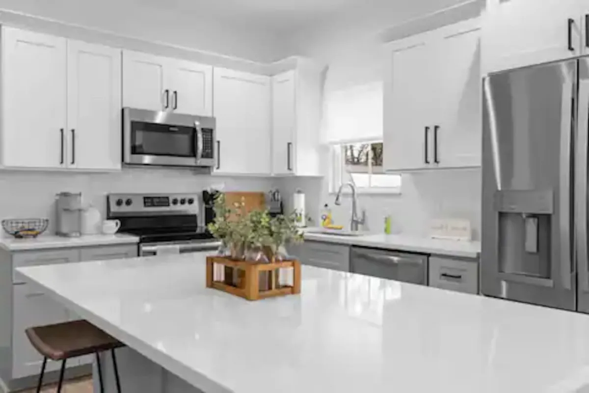 A bright and contemporary kitchen featuring a large island, stainless steel appliances, white cabinetry, and a floral centerpiece. The space is open and inviting, with plenty of counter space for meal prep or group cooking. Bar stools at the island suggest room for casual dining or collaborative culinary efforts.