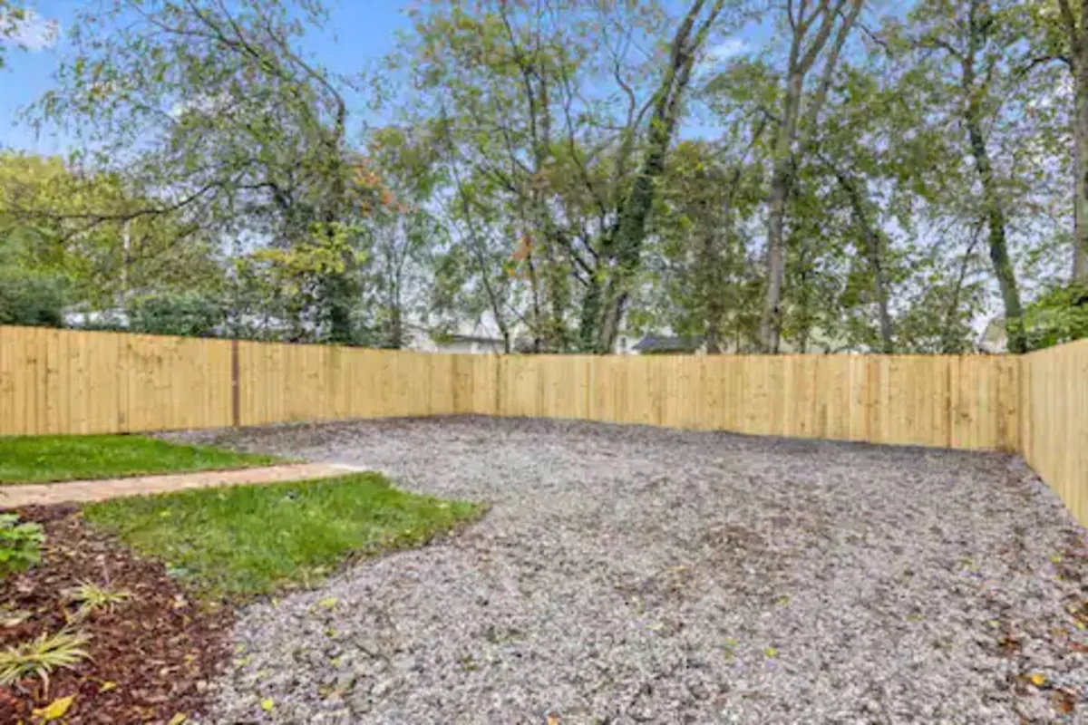 This image features a spacious backyard with a gravel surface, neatly defined green grass, a wooden fence, and a line of mature trees in the background. The open space is uncluttered, providing ample room for outdoor games, gatherings, and relaxation.