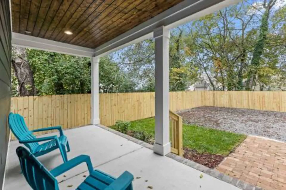 The photo depicts a cozy outdoor entertaining space with several blue Adirondack chairs arranged on a patio under a wooden ceiling, all set within a fenced backyard. The area is ideal for relaxing, socializing, or hosting a barbecue.