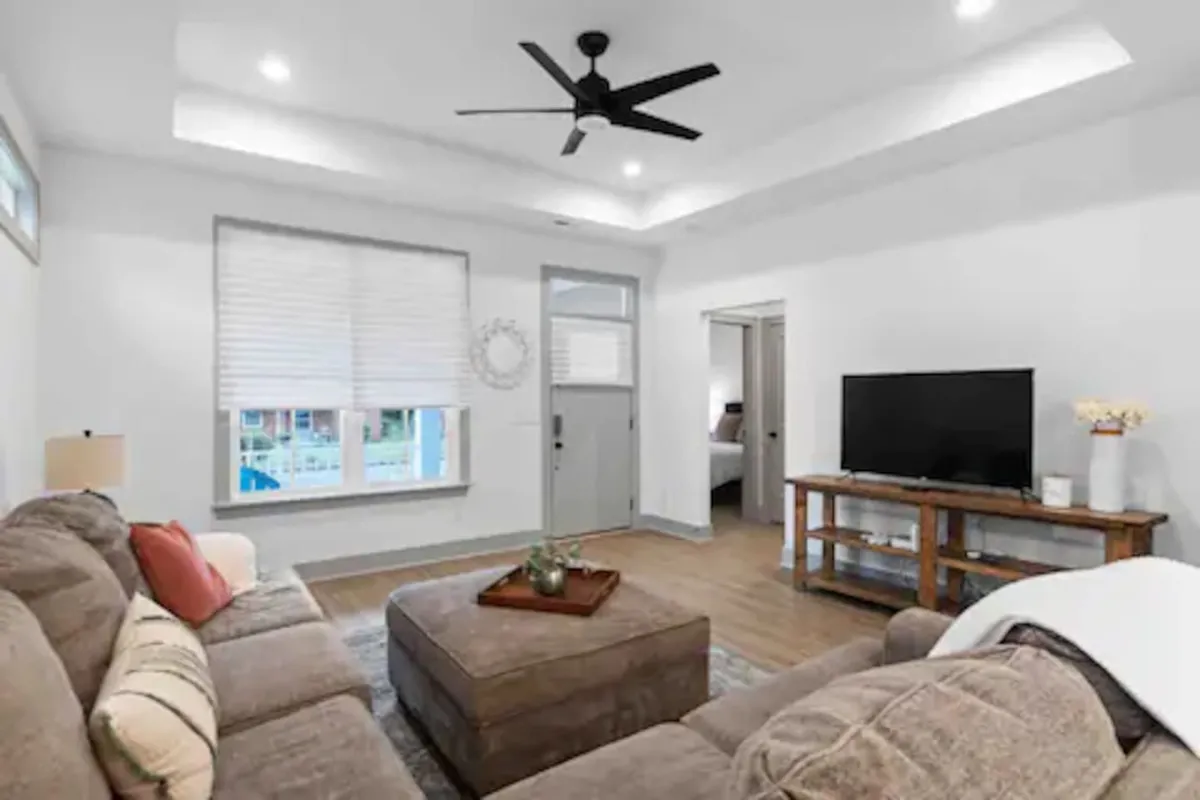 The image showcases a cozy, inviting living room with a comfortable sofa, stylish coffee table, large TV, and plenty of natural light streaming in from a window beside the door. The seating arrangement invites relaxation and connection, perfect for gatherings or movie nights with friends and family.