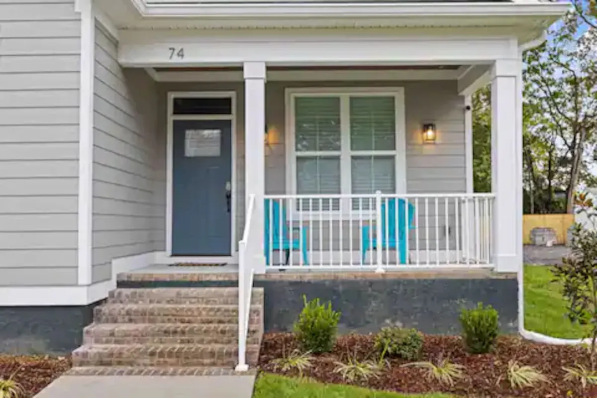 An exterior perspective of a front porch with blue chairs and a front door, providing a view that connects the home to the surrounding neighborhood. The porch looks inviting and is framed by gentle light, hinting at a relaxed, social atmosphere.