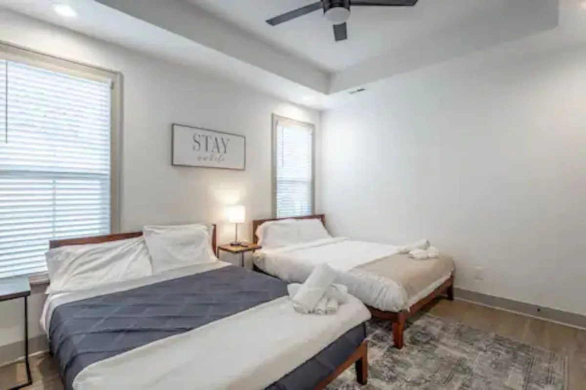 A comfortable bedroom containing two beds, each with crisp linens and decorative pillows. A nightstand with a lamp sits between the beds, and the wall above is adorned with subtle artwork. The space is well-lit and designed for both relaxation and quiet conversation, accommodating multiple guests with ease.